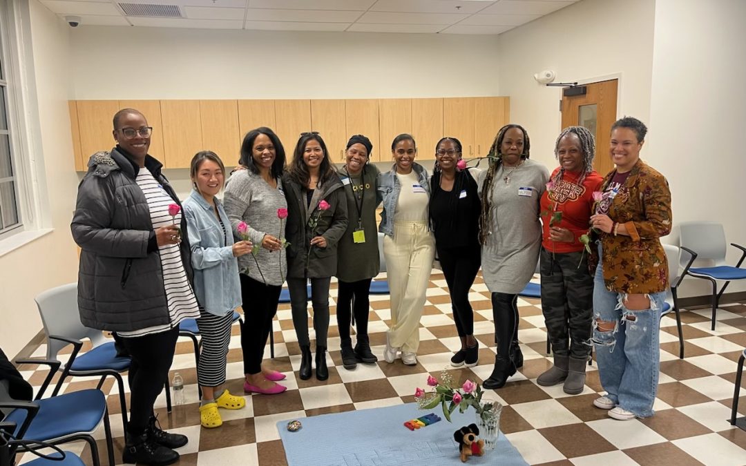 a group of women smile in a circle while holoding flowers, a small cloth with a bouquet of flowers sits in the center of the group.